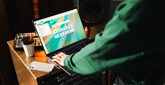 Person browsing Opera's Start page on a laptop in a home music studio, with a coffee and phone nearby.
